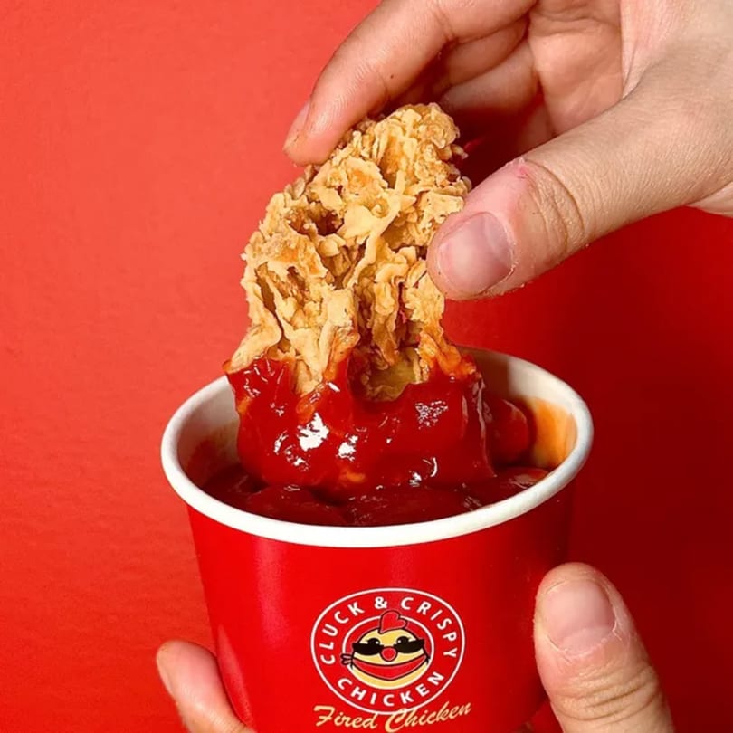 A hand dips a crispy fried chicken piece into a red cup of Cluck & Crispy sauce.