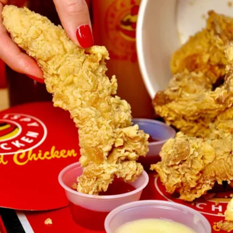 A hand with red nail polish dips a crispy chicken tender into a small cup of red sauce.