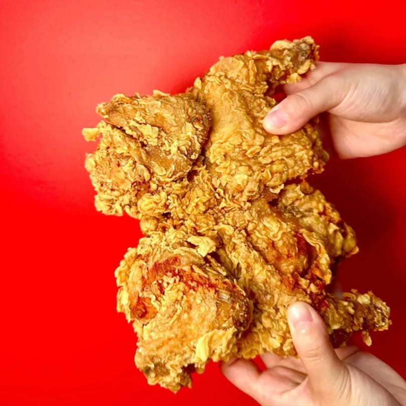 Two hands holding a large piece of crispy fried chicken against a vibrant red background.
