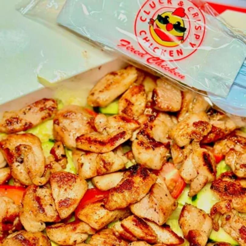 Grilled chicken pieces over a salad with lettuce, tomatoes, and cucumbers, with a Cluck & Crispy bag.