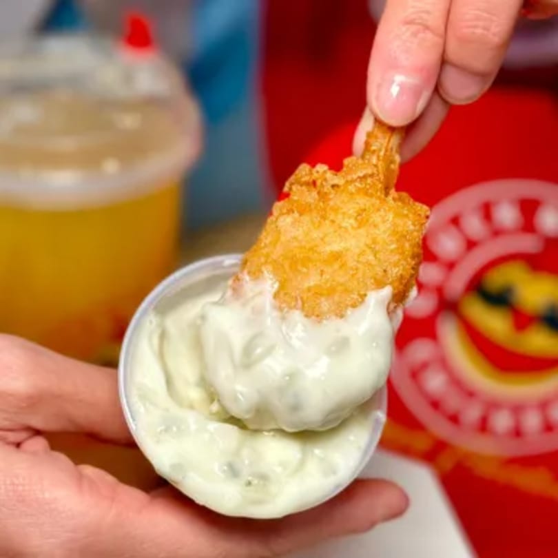 A hand dips a crispy fried chicken tender into a small cup of white sauce.