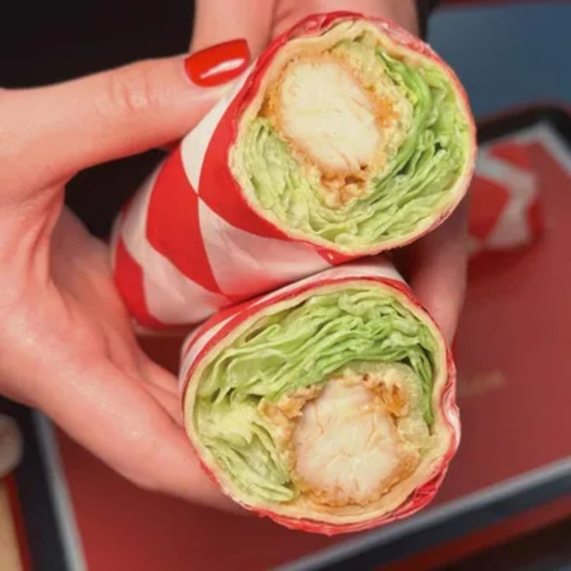 Two hands holding a Cluck & Crispy chicken wrap, cut in half, showing crispy chicken and lettuce.