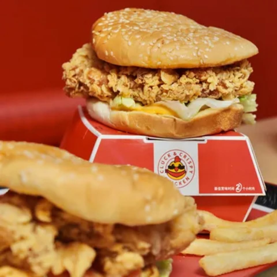 Two crispy chicken sandwiches with fries, one on a Cluck & Crispy box, against a red background.