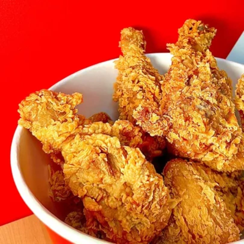 A white bucket filled with golden-brown, crispy fried chicken pieces against a red background.