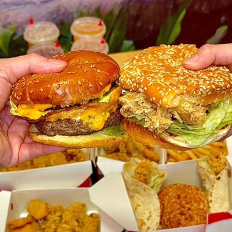 Hands holding a beef burger and a crispy chicken burger, with sides in boxes below.