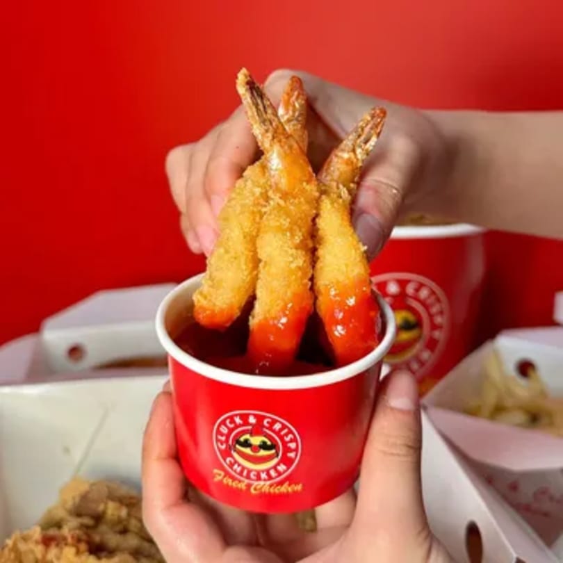Three crispy fried shrimp being dipped into a red cup of sauce from Cluck & Crispy Chicken.
